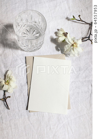 Wedding stationery set. Mock-up scene with blank paper greeting cards, envelope and glass of water on linen tablecloth background. White magnolia stellata tree branches in sunlight.Vertical still life Wedding stationery set. Mock-up scene with blank paper greeting cards, envelope and glass of water on linen tablecloth background. White magnolia stellata tree branches in sunlight.Vertical still life 64557953