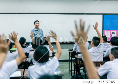 A smiling Asian male high school teacher teaches the white uniform students in the classroom by asking questions and then the students raise their hands for answers. 64597385