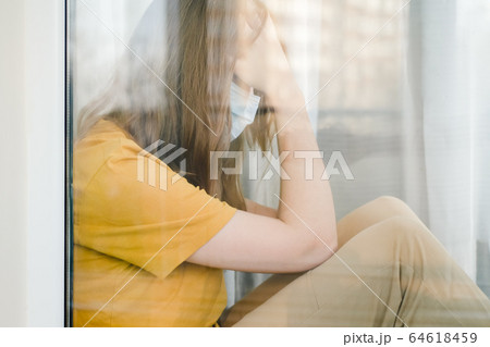 Sad girl in medical protective mask and yellow t-shirt sitting on the windowsill during a pandemic quarantine coronavirus Covid-19. Isolation at home. Sad girl in medical protective mask and yellow t-shirt sitting on the windowsill during a pandemic quarantine coronavirus Covid-19. Isolation at home. 64618459