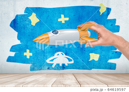 Man's hand playing with silver and orange space rocket against wall with sketchy painting of blue starry sky and small UFO in it. 64619597