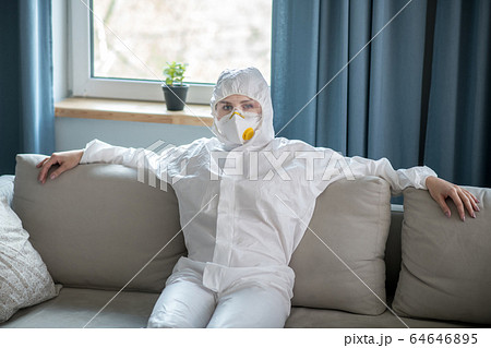 Woman in white workwear and respirator sitting on the sofa 64646895