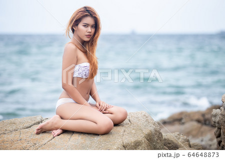 Beautiful girl blonde in white bikini seating on the rock. 64648873