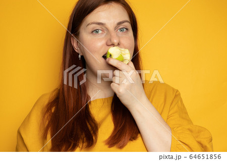 Proper nutrition balance concept. Young girl eats a green apple, standing on a yellow wall background Proper nutrition balance concept. Young girl eats a green apple, standing on a yellow wall background 64651856