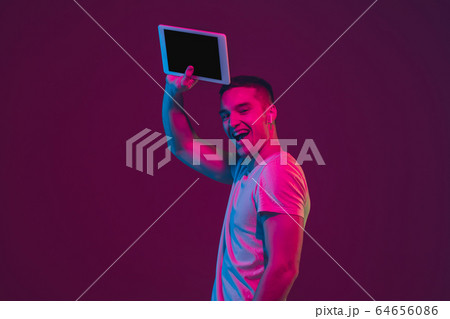 Caucasian man's portrait isolated on pink-purple studio background in neon light, using tablet Caucasian man's portrait isolated on pink-purple studio background in neon light, using tablet 64656086
