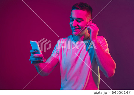 Caucasian man's portrait isolated on pink-purple studio background in neon light, with smartphone and earphones Caucasian man's portrait isolated on pink-purple studio background in neon light, with smartphone and earphones 64656147