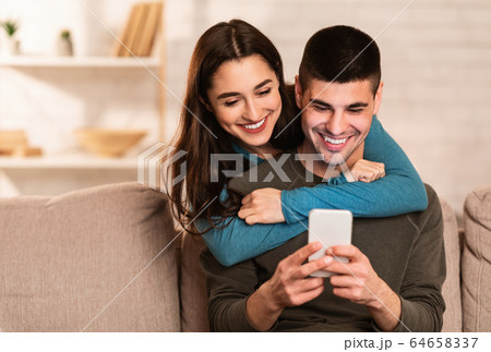 Lovely couple using cell phone sitting on sofa Lovely couple using cell phone sitting on sofa 64658337