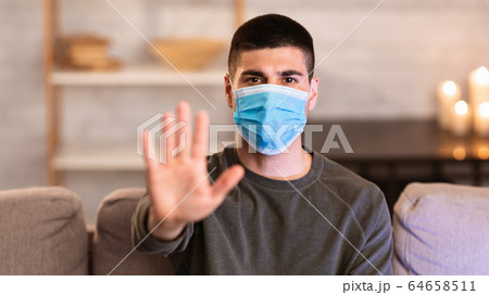 Serious guy in face mask gesturing stop sitting on couch 64658511