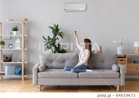 Young woman switching on air conditioner sitting on couch 64659673