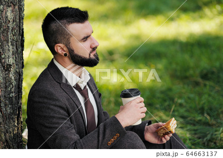 Young attractive bearded business man sitting on green grass under tree and resting in park. Drink coffee and eat sandwich. Relaxed, tired of work, lunch break. Young attractive bearded business man sitting on green grass under tree and resting in park. Drink coffee and eat sandwich. Relaxed, tired of work, lunch break. 64663137