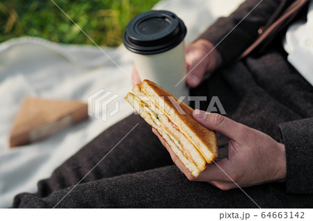 Hands of a man in a suit with a glass of takeaway coffee and a triangular sandwich with tuna. 64663142
