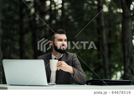 Young attractive business man in suit and tie sits at desk and works on computer outdoors. Drink coffee from white cup. Green trees, nature and park on background. 64678319