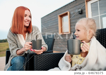 A young mother and little daughter sit in chairs in warm rugs and drink tea from the mugs on the terrace of a wooden house. Autumn evening in the country. A young mother and little daughter sit in chairs in warm rugs and drink tea from the mugs on the terrace of a wooden house. Autumn evening in the country. 64678375