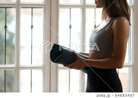 Female wearing sportswear standing near window holding yoga mat 64683497