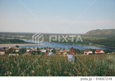 a little girl in a loose dress with loose hair walks in a meadow. Village houses, forest and river as background. The concept of summer, warmth, freedom, village life, sunburn, chldhood 64687248