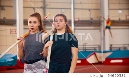 Pole vault training - two women standing on the stadium holding their poles and looking in the camera Pole vault training - two women standing on the stadium holding their poles and looking in the camera 64690581