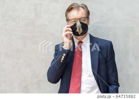 Mature businessman wearing mask and talking on the phone against concrete wall Mature businessman wearing mask and talking on the phone against concrete wall 64715862