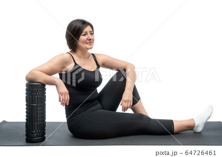A middle-aged woman, on a gymnastic mat with myofascial roller, is resting after exercise on a white background. A middle-aged woman, on a gymnastic mat with myofascial roller, is resting after exercise on a white background. 64726461
