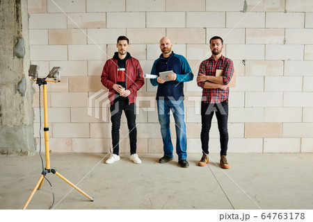 Three young male engineers or builders in workwear standing in line along wall Three young male engineers or builders in workwear standing in line along wall 64763178