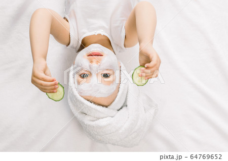 Funny little girl with a mask for skin face and towel on her head, holding two slices of fresh cucumber and looking at camera, while lying on white background after bath 64769652