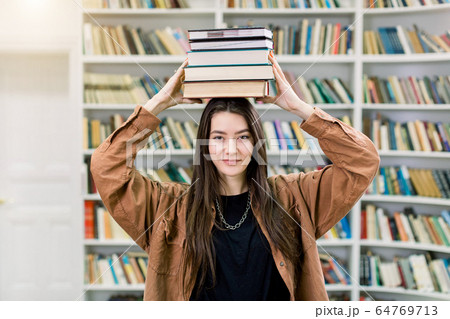 Female student balancing books on her head, standing in college library and looking at camera. Beautiful girl holds a stack of books on her head and smiles. Education concept, studying with books 64769713