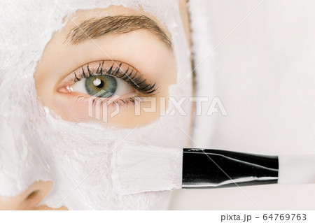 Face mask, skin care, beauty concept. Cropped close up macro photo of beautiful young girl at spa, and cosmetician brush applying clay facial mask. Copy space, isolated on white background. Face mask, skin care, beauty concept. Cropped close up macro photo of beautiful young girl at spa, and cosmetician brush applying clay facial mask. Copy space, isolated on white background. 64769763