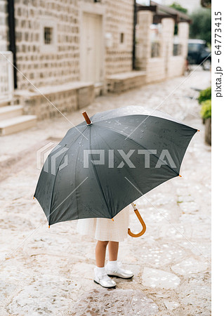 The child poses with an umbrella. A little girl in a dress stands outside under an umbrella black during the rain. 64773415
