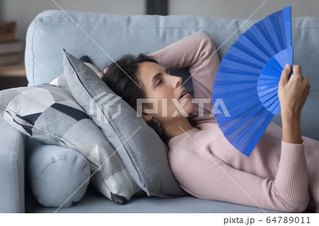 Unhealthy millennial girl cooling herself with paper fun indoors. Unhealthy millennial girl cooling herself with paper fun indoors. 64789011