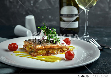 White steak of grilled fish on pillow of vegetables and yellow sauce, with tomatoes and fresh herbs. Fish on white plate with white wine on dark beautiful background. Close-up. Space 64793098