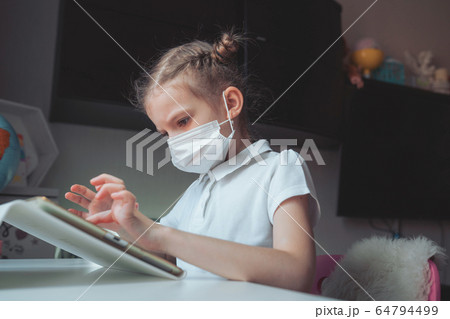 Caucasian preteen girl with medical mask on her face concentrated on her task with tablet. Concept of distance learning in isolation while coronavirus Caucasian preteen girl with medical mask on her face concentrated on her task with tablet. Concept of distance learning in isolation while coronavirus 64794499