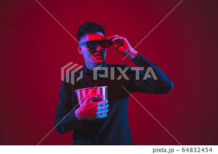 Caucasian man's portrait isolated on red studio background in purple-blue neon light, emotional and beautiful, looking throught 3D-eyewear, cinema 64832454