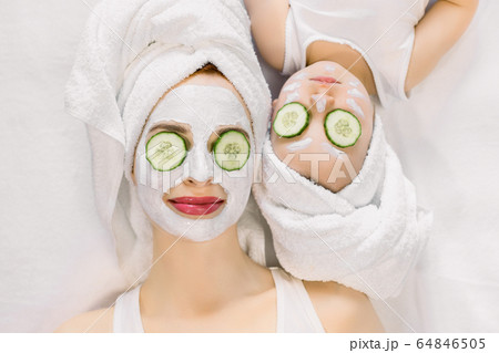 Mother and little daughter having spa procedures together. They are in white bath towels on head and with slices of cucumber on their eyes. Woman has white facial mask on her skin 64846505