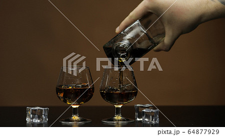 Pouring whiskey, cognac into glass. Brown background. Pour of alcohol drink 64877929