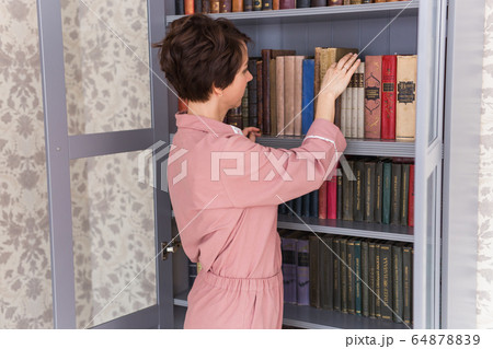 Young caucasian young woman with brunette hair taking boor for reading on free time standing near bookcase, positive girl selecting literature taking novels from shelves in library. Stay home. 64878839