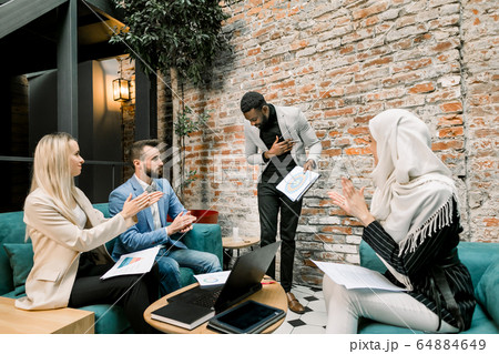 Leadership and business speech. Handsome young African man in grey jacket finished his successful speech or report for multiethnic young colleagues, sitting on sofas and clapping hands 64884649