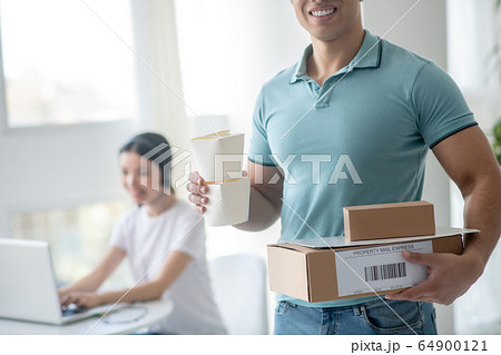 Male courier holding food boxes and parcels, dark-haired female typing on laptop behind him 64900121