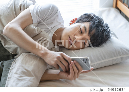 ベッドに寝ながらスマートフォンを見る若い男性 64912634