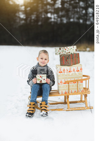 Cute little child, boy, sitting on a wooden sledge decorated with presents boxes, holding the box with small gift, outdoors in winter forest or park, snow wintertime 64918210