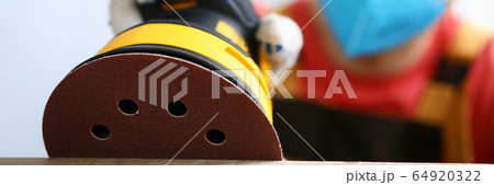 Masked carpenter grinds wood with sander machine 64920322
