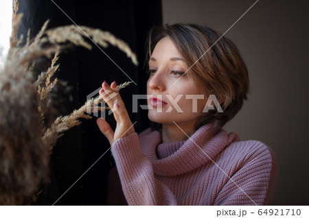 Portrait of a young beautiful woman touching wheat spikelets. Blonde girl wearing pink sweater in scandinavian interior. Atmospheric hygge style Portrait of a young beautiful woman touching wheat spikelets. Blonde girl wearing pink sweater in scandinavian interior. Atmospheric hygge style 64921710
