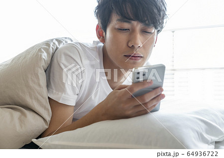 布団の中でスマホを見る若い男性 64936722
