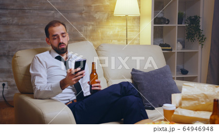 Tired businessman sitting on couch scrolling on his phone Tired businessman sitting on couch scrolling on his phone 64946948