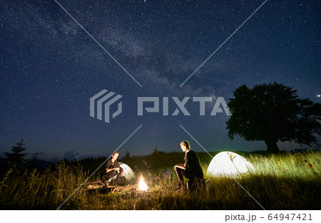 Friends travellers spending time in the mountains sitting around bonfire in camping under beautiful starry sky 64947421
