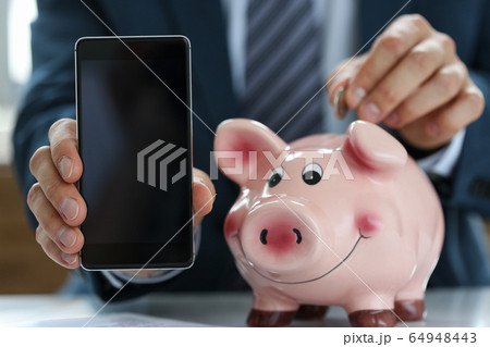 In hands smartphone, man puts coin in piggy bank 64948443