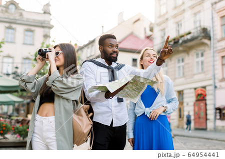 Multi-ethnic travelers using local map together on sunny day, African man pointing forward and showing something to blond girl. Pretty brunette Caucasian woman makes photos on her camera Multi-ethnic travelers using local map together on sunny day, African man pointing forward and showing something to blond girl. Pretty brunette Caucasian woman makes photos on her camera 64954441