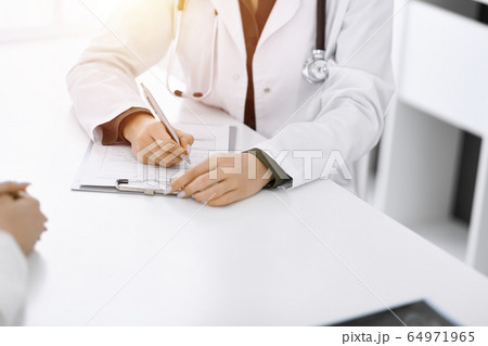 Unknown woman-doctor and female patient sitting and talking at medical examination in sunny clinic, close-up. Therapist filling up medication history record. Medicine concept Unknown woman-doctor and female patient sitting and talking at medical examination in sunny clinic, close-up. Therapist filling up medication history record. Medicine concept 64971965