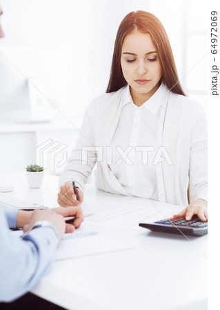 Young business woman and man sitting and working with computer and calculator in sunny office. Bookkeeper checking balance. Tax and audit concepts 64972069