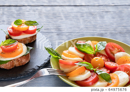 Bruschetta and salad Caprese: red and yellow tomatoes, mozzarella cheese, green Basil, herbs, olive oil on shale board and grey wooden table with copy space. Selective focus. Bruschetta and salad Caprese: red and yellow tomatoes, mozzarella cheese, green Basil, herbs, olive oil on shale board and grey wooden table with copy space. Selective focus. 64975676