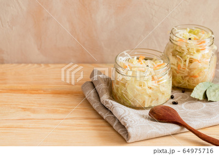 Two glass jars with sauerkraut and wooden spoon and spices is standing on cooking towel on wooden table on light background with copy space. Healthy fermented food. Two glass jars with sauerkraut and wooden spoon and spices is standing on cooking towel on wooden table on light background with copy space. Healthy fermented food. 64975716