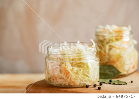 Two glass jars with sauerkraut are standing on wooden cooking board on light background with copy space. Healthy fermented food. Selective focus. Two glass jars with sauerkraut are standing on wooden cooking board on light background with copy space. Healthy fermented food. Selective focus. 64975717