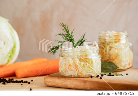 Glass jars with sauerkraut are standing on wooden cooking board and ingredients for its preparation: cabbage and carrots on light background. Glass jars with sauerkraut are standing on wooden cooking board and ingredients for its preparation: cabbage and carrots on light background. 64975718